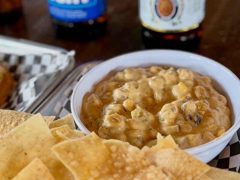 Tortilla chips and a bowl of warm, chunky corn dip with bottled beers behind
